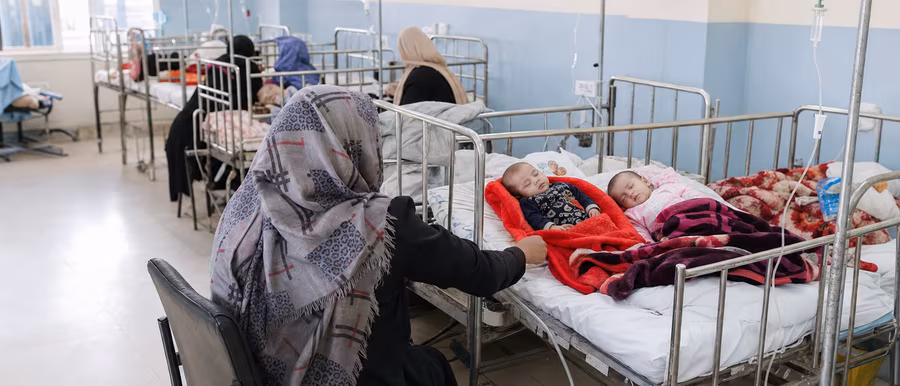 Two babies sharing a single hospital crib bed in a crowded pediatric ward, with a mother attending to them beside IV stands