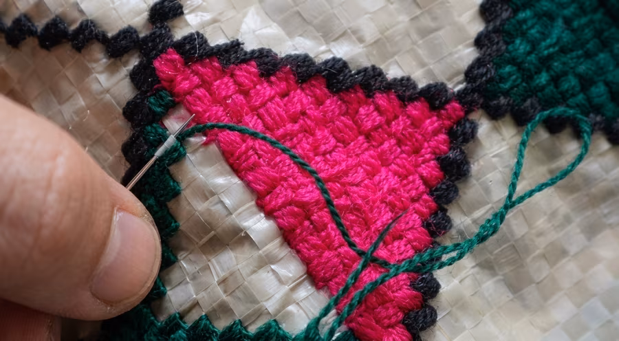 A hand guiding a needle and green thread through a pink and black cross-stitch pattern on woven sack fabric