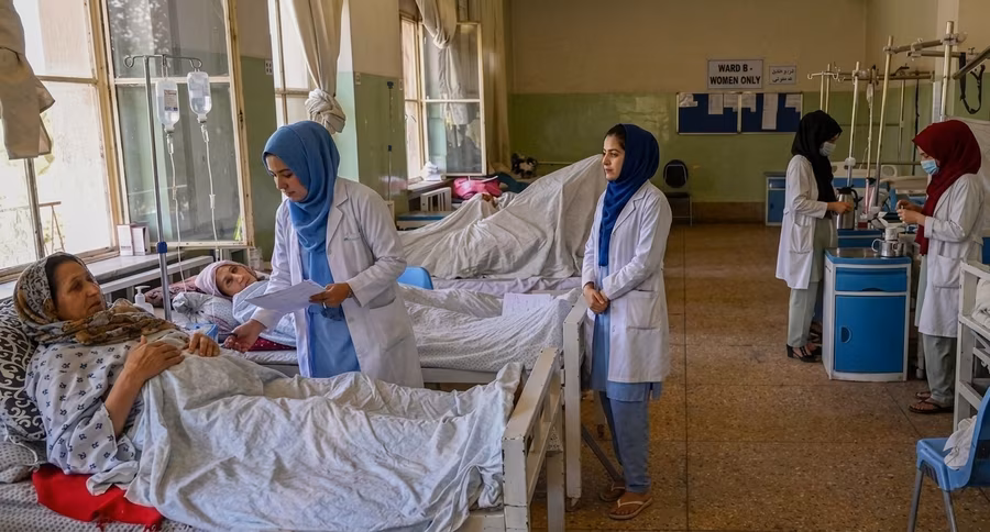 Female doctors in white coats checking on patients in a women&rsquo;s hospital ward, with IV drips beside the beds