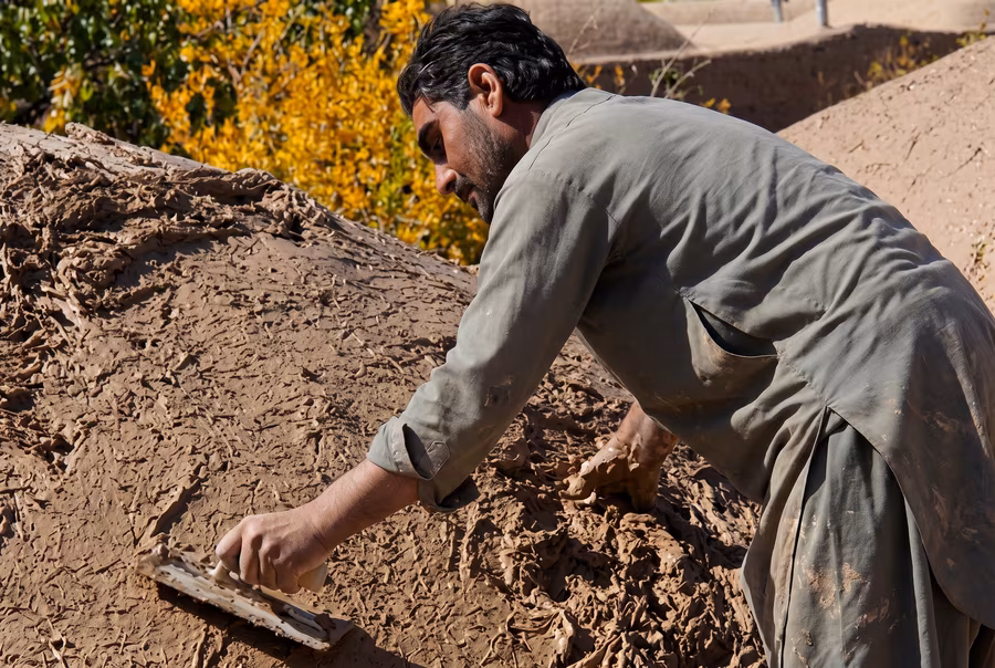 A man kneeling on a mound of wet clay and straw, smoothing it with a flat hand trowel