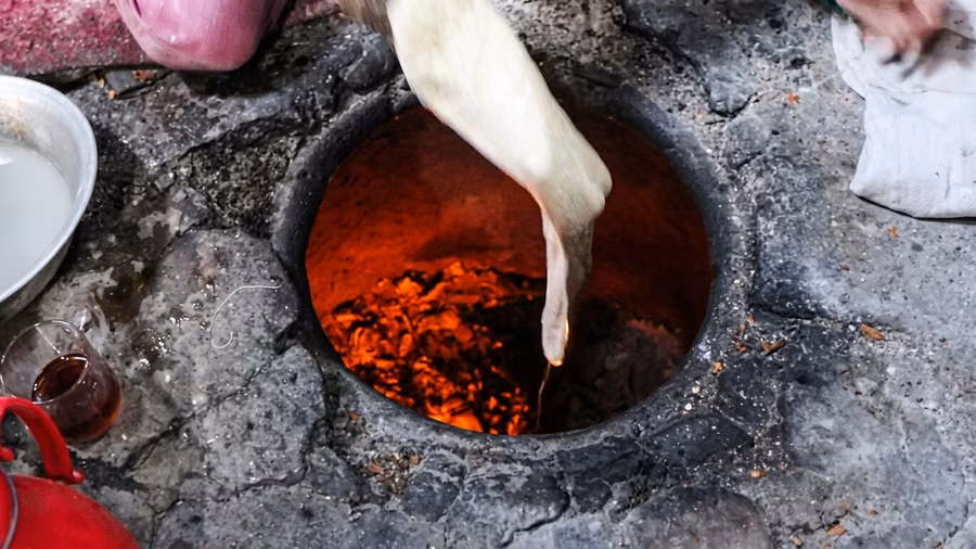 A stretched piece of dough being lowered into a tandoor pit, its tip nearly touching the glowing coals below