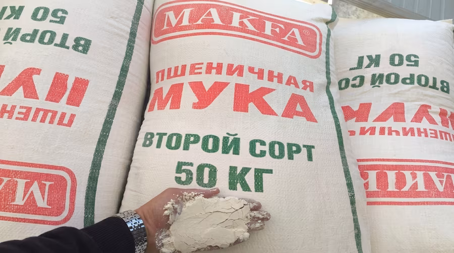 A hand holding a scoop of flour in front of stacked imported Kazakh flour sacks