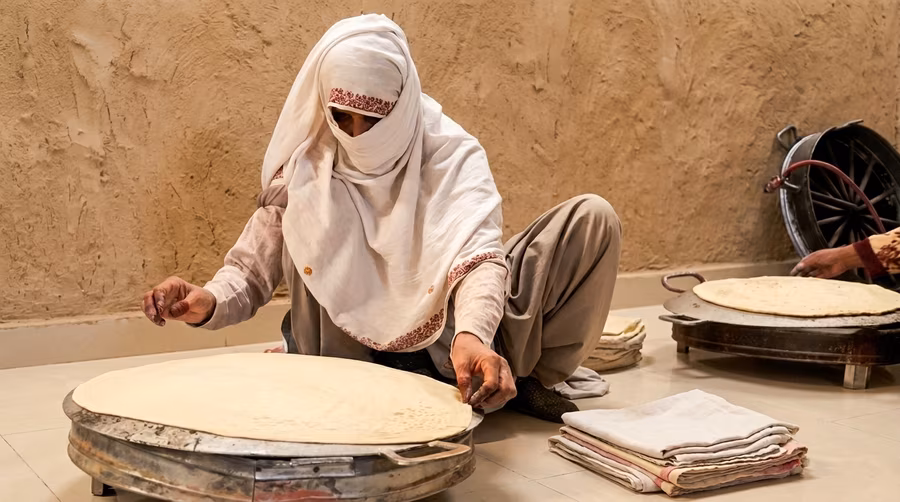 A woman shaping chapati dough on a griddle, with another loaf already cooked beside her