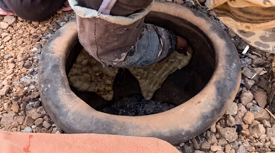 Bread being pressed down into a clay tandoor set low into the ground, glowing coals beneath
