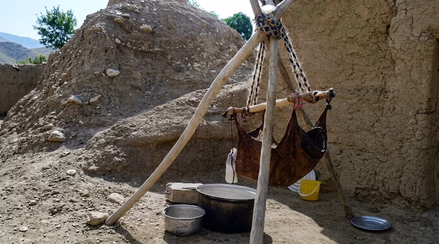 A worn leather churn hanging from a wooden tripod, with pots and a yellow bucket set on the ground below it