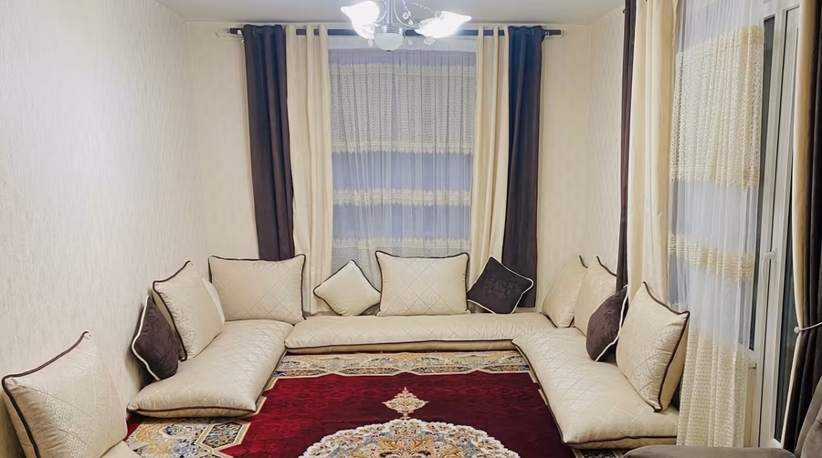 A neatly arranged Afghan guest room with cream-colored toshak and cushions placed around the walls, over a patterned carpet